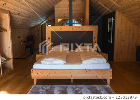 A cozy hotel room with natural wood paneling, a large wooden bed, built in nightstands, a sloped ceiling, and natural light in the Italian Alps. 125012979