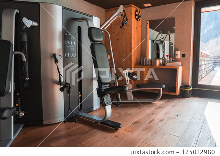 A gym with wooden interiors, Technogym equipment, and a balcony overlooking snow covered peaks in a luxury hotel in the Italian Alps. 125012980