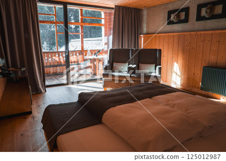 A well lit room with wooden paneling, a large bed, armchairs, and a balcony overlooking a snow covered roof and forested area in the Italian Alps. 125012987