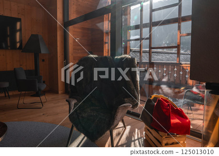Cozy interior with wooden walls, fur draped chair, sunlit balcony, and snow covered peaks visible through a large glass window. 125013010