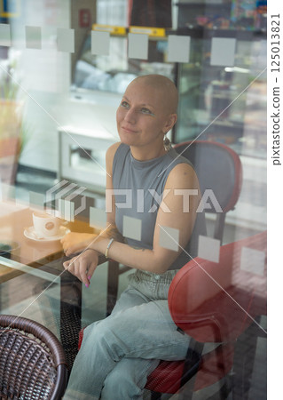 Young woman with cancer sitting inside a cafe, seen through the window, lost in thought. The concept highlights how people with serious illnesses continue to live actively and be part of society 125013821
