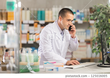 Male pharmacist is talking on mobile phone with patient - checking data on computer Male pharmacist is talking on mobile phone with patient - checking data on computer 125016005