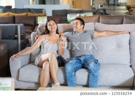 In store,husband and wife are sitting on gray sofa and choose upholstered furniture for living room 125016254