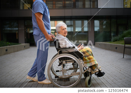 Side view of caregiver pushing senior woman in wheelchair. 125016474