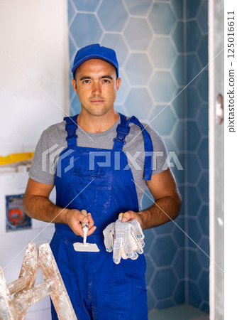 Professional man builder in blue construction overalls and cap posing with gloves and trowel in apartment 125016611