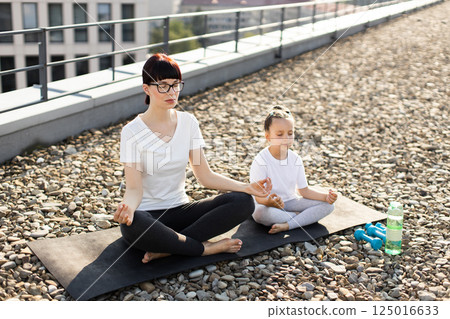 Woman and girl meditating together in lotus position on rooftop, enjoying outdoor relaxation. Woman and girl meditating together in lotus position on rooftop, enjoying outdoor relaxation. 125016633