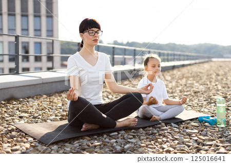 Mother and daughter practicing yoga on rooftop. Young woman and girl meditating outdoors, serene atmosphere. Family bond, yoga lifestyle, healthy living. Mother and daughter practicing yoga on rooftop. Young woman and girl meditating outdoors, serene atmosphere. Family bond, yoga lifestyle, healthy living. 125016641