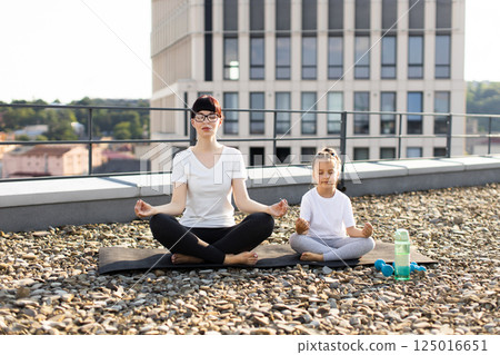 Woman and young girl practicing meditation outdoors on urban rooftop, expressing mindfulness and tranquility. Woman and young girl practicing meditation outdoors on urban rooftop, expressing mindfulness and tranquility. 125016651
