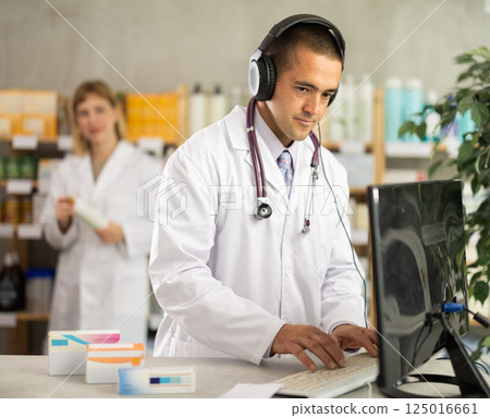 Male pharmacist with headphones working with computer while female pharmacist standing in background 125016661