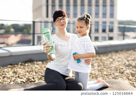 Woman and young girl sitting on rooftop exercising together holding green water bottle, smiling Woman and young girl sitting on rooftop exercising together holding green water bottle, smiling 125016664