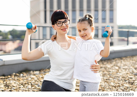 Mother and young daughter with dumbbells rooftop cityscape exercising smiling sunny day outdoor workout inspiration Mother and young daughter with dumbbells rooftop cityscape exercising smiling sunny day outdoor workout inspiration 125016669