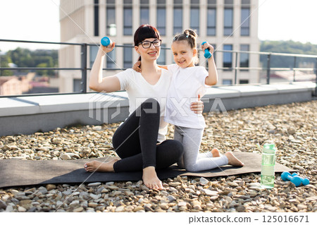 Mother with daughter exercising together using dumbbells on rooftop, engaging in outdoor fitness. Models are female, mother is adult, daughter is child. 125016671