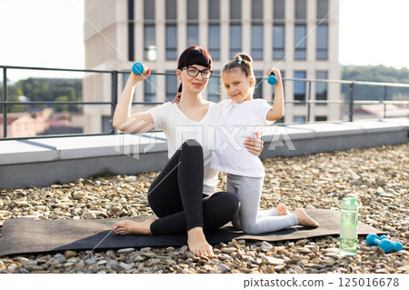 Young adult woman and daughter practicing fitness with dumbbells outdoors on rooftop terrace, showcasing healthy lifestyle concept. 125016678