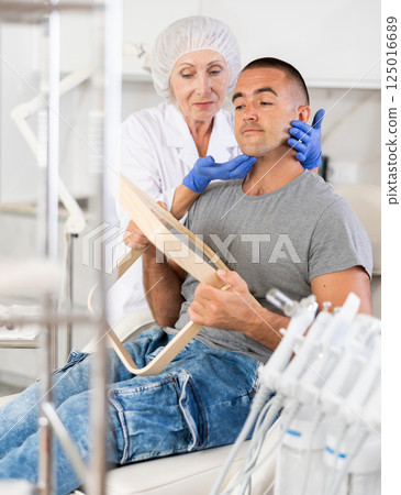 Portrait of man sitting and looking at mirror during appointment with doctor at cosmetology clinic Portrait of man sitting and looking at mirror during appointment with doctor at cosmetology clinic 125016689