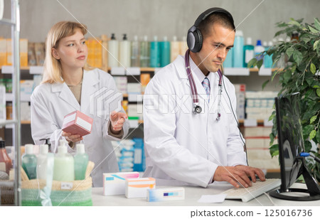 Man pharmacist works at computer using headset, girl assistant in background Man pharmacist works at computer using headset, girl assistant in background 125016736