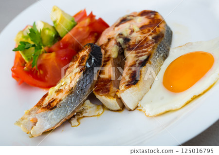 Grilled salmon with fried egg, tomatoes, avocado on white dish 125016795