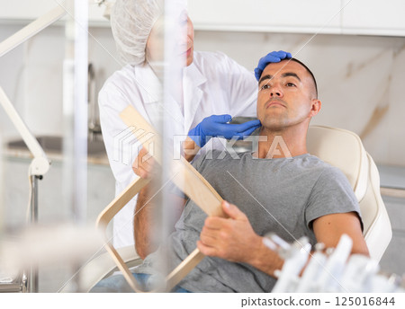 Female doctor, together with male patient, examines face in mirror after undergoing hardware procedure for skin rejuvenation Female doctor, together with male patient, examines face in mirror after undergoing hardware procedure for skin rejuvenation 125016844