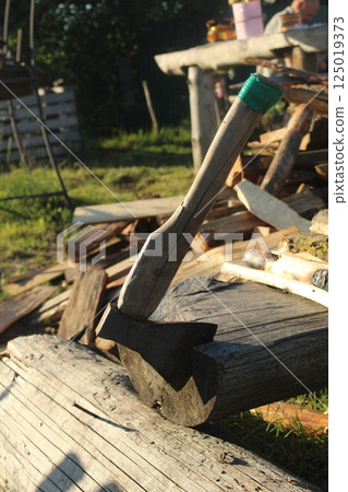 The axe sticks out of a large log against the background of other firewood. Harvesting firewood by hand with an axe The axe sticks out of a large log against the background of other firewood. Harvesting firewood by hand with an axe 125019373