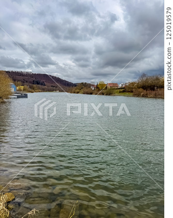 Tranquil river with cloudy sky and hills in distance, rural village shoreline. High quality photo 125019579