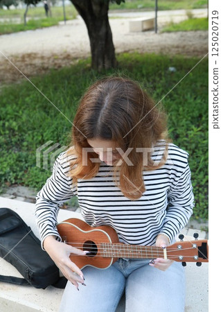 Young Woman Tunes The Ukulele Musical Instrument In A Park Outside 125020719