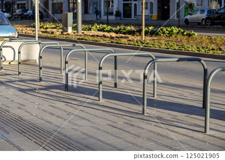 Empty metal bike racks casting long shadows on sunlit city sidewalk near road and plant beds 125021905