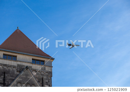 Airplane flying over historic stone building with red tiled roof against bright blue sky 125021909