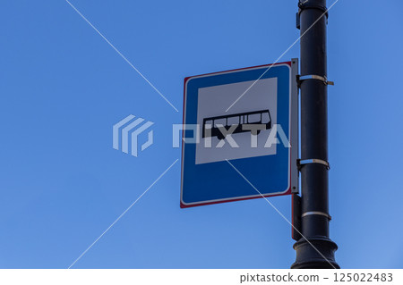 Public bus stop sign mounted on black pole against clear blue sky in urban environment, minimal composition Public bus stop sign mounted on black pole against clear blue sky in urban environment, minimal composition 125022483