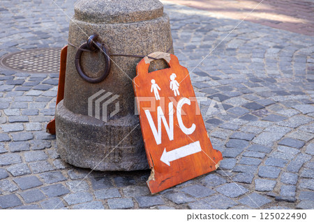 Handmade wooden WC sign with male and female icons and arrow leaning against stone post on old cobblestone street Handmade wooden WC sign with male and female icons and arrow leaning against stone post on old cobblestone street 125022490