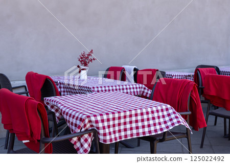 Outdoor cafe tables with red and white checkered tablecloths and blankets on chairs against plain wall on quiet city street Outdoor cafe tables with red and white checkered tablecloths and blankets on chairs against plain wall on quiet city street 125022492