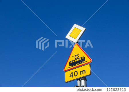 Urban traffic signs with tramway warning and priority road indicator against clear blue sky on sunny day, close-up view from below 125022513