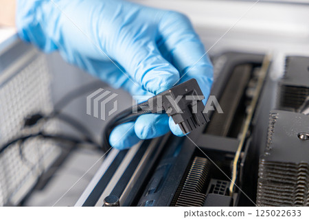 Close-up of technician hand in blue glove holding GPU 90-degree power connector above graphics card during computer assembly process 125022633