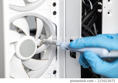 Technician in blue glove using soft brush to remove dust from dirty cooling fan inside computer case during PC cleaning 125022672