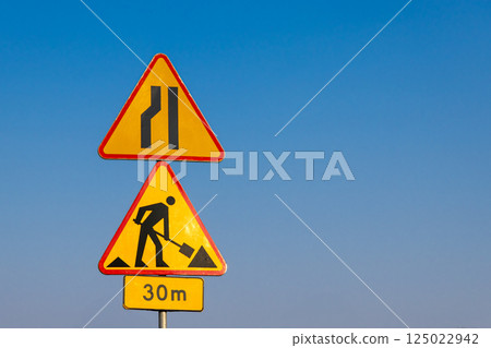 Road construction and lane reduction warning signs against clear blue sky indicating roadwork ahead in 30 meters distance on highway or city road 125022942