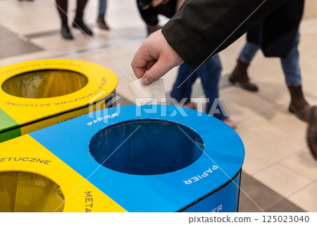 Person throwing paper receipt into blue recycling bin labeled papier in shopping mall, with colorful containers for plastic and metal in background Person throwing paper receipt into blue recycling bin labeled papier in shopping mall, with colorful containers for plastic and metal in background 125023040