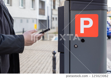 Man using smartphone near solar-powered parking meter on urban street, making mobile payment for parking spot in city area, close-up of hand and modern machine 125023062