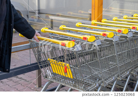 Row of shopping carts with yellow handles connected by coin chain locks near glass wall outside supermarket entrance with hand grabbing one trolley Row of shopping carts with yellow handles connected by coin chain locks near glass wall outside supermarket entrance with hand grabbing one trolley 125023068