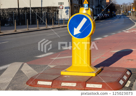 Directional road sign with white arrow on yellow post indicating traffic must pass on the left, standing on pedestrian island in urban street under sunlight 125023073