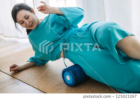 Woman rolling hips on a blue foam roller on cork mat at home. Myofascial release, body awareness, mobility training, restorative wellness Woman rolling hips on a blue foam roller on cork mat at home. Myofascial release, body awareness, mobility training, restorative wellness 125023267