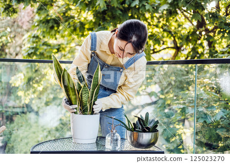 Young woman bending over a potted plant as she repots it on a sunlit balcony. DIY gardening, indoor plant care, self-reliant lifestyle, hands-on wellness 125023270