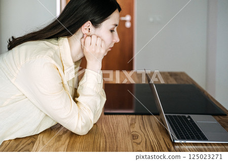 Young Asian woman sitting at a wooden table, staring at a laptop screen with a serious expression. Job insecurity, AI disruption, remote work stress, automation impact 125023271