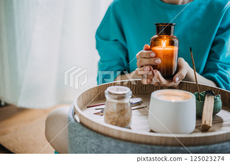 Close-up of hands holding a lit amber candle above a tray with incense, palo santo, and matches. Ritual details of modern mindfulness, intentional objects, palo santo practice, sensory wellness Close-up of hands holding a lit amber candle above a tray with incense, palo santo, and matches. Ritual details of modern mindfulness, intentional objects, palo santo practice, sensory wellness 125023274