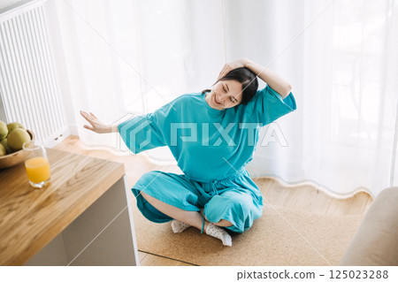Young white woman in a turquoise robe does a neck stretch while sitting cross-legged on a mat in a bright room. Soft wellness, mindful morning routine, home self-care, body awareness Young white woman in a turquoise robe does a neck stretch while sitting cross-legged on a mat in a bright room. Soft wellness, mindful morning routine, home self-care, body awareness 125023288