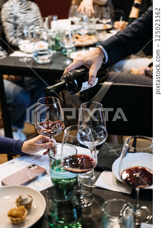 Pouring wine into glasses at a formal group tasting event. Community rituals, curated hospitality, beverage storytelling Pouring wine into glasses at a formal group tasting event. Community rituals, curated hospitality, beverage storytelling 125023362
