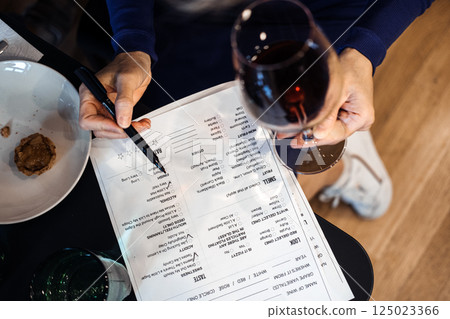 Close-up of a person holding a glass of red wine while writing on a wine workshop menu. Sommelier training, wine tasting analysis, hands-on learning, sensory evaluation 125023366