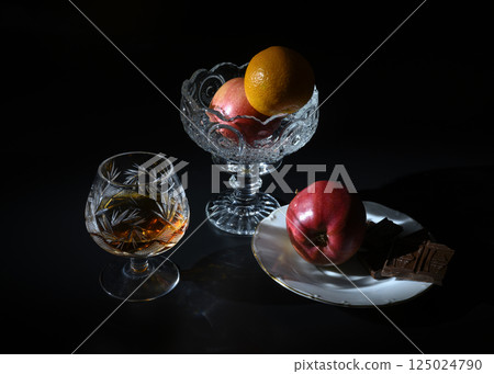 A crystal glass with brandy, a bowl with apple and tangerine, and chocolate A crystal glass with brandy, a bowl with apple and tangerine, and chocolate 125024790