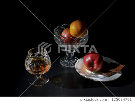A crystal glass with brandy, a bowl with apple and tangerine, and chocolate A crystal glass with brandy, a bowl with apple and tangerine, and chocolate 125024791