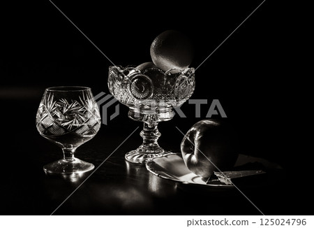 A crystal glass with brandy, a bowl with apple and tangerine, and chocolate, in black and white A crystal glass with brandy, a bowl with apple and tangerine, and chocolate, in black and white 125024796
