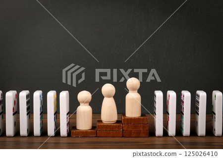 Wooden Figures Standing on Stacked Blocks with Dominoes in Background 125026410