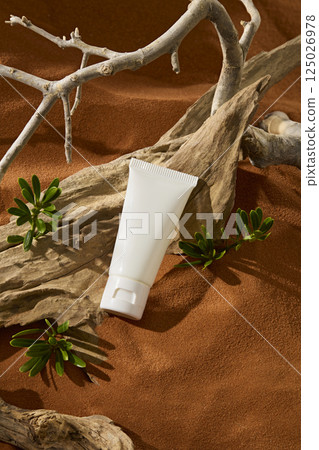 White tube without label placed on a rough and curvy wooden trunk. Grey dry branches with desert shrub growing around them. Fine brown sand undulating in natural ripples. Vertical photo.  125026978