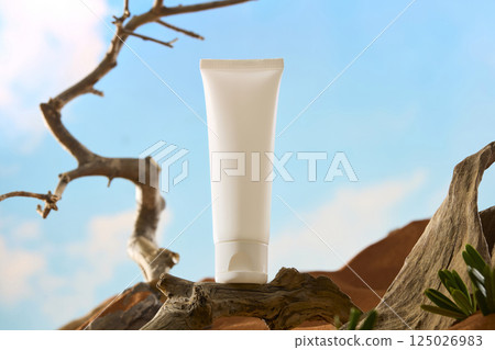 Mockup of cosmetic tube placed on horizontal tree trunks. The tree trunk is curved and has several sharp thorns. The desert is hot and barren with white clouds and bright sunlight in the sky. 125026983